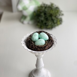 May include: A white ceramic pedestal with a nest of twigs and three light blue Easter eggs.