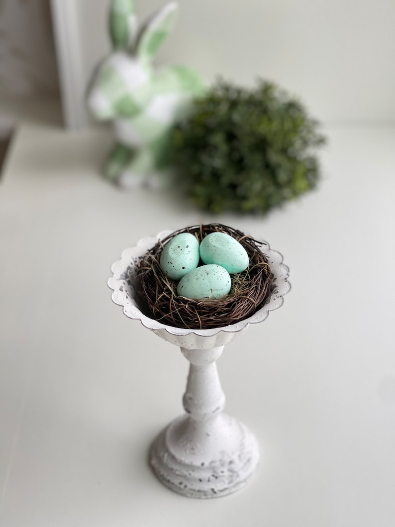 May include: A white ceramic pedestal with a nest of twigs and three light blue Easter eggs.