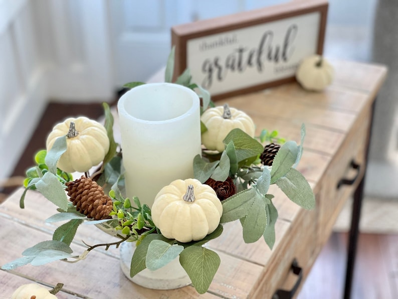 May include: A white candle in a glass jar surrounded by a wreath of white pumpkins, pine cones, and green leaves. The wreath is sitting on a wooden table with a sign that says "thankful grateful" in the background.