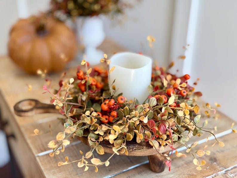 May include: A wooden pedestal with a rustic finish holds a fall-themed wreath. The wreath is made of faux greenery with small orange berries and a white pillar candle in the center.