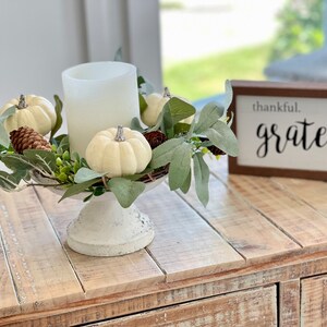May include: White pillar candle with a white frosted glass holder, surrounded by a wreath of green leaves and white pumpkins. The wreath is on a white pedestal. A wooden sign with the text "thankful. grateful bless" is in the background.