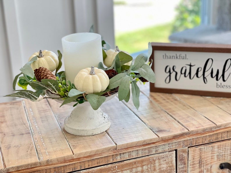 May include: White pillar candle with a white frosted glass holder, surrounded by a wreath of green leaves and white pumpkins. The wreath is on a white pedestal. A wooden sign with the text "thankful. grateful bless" is in the background.