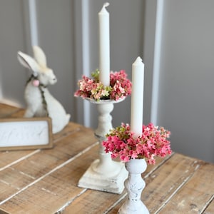 May include: Two white candle holders with pink flowers around the base and white candles. The candle holders are on a wooden table.