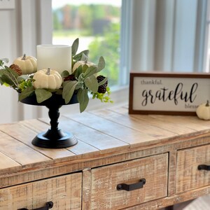 May include: A white candle sits in a black pedestal stand surrounded by a wreath of white pumpkins, green leaves, and pine cones. A wooden sign with the text "thankful. grateful. bless." sits on a light wood table with two drawers.