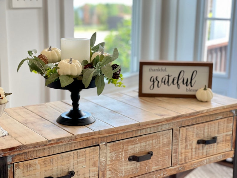 May include: A white candle sits in a black pedestal stand surrounded by a wreath of white pumpkins, green leaves, and pine cones. A wooden sign with the text "thankful. grateful. bless." sits on a light wood table with two drawers.