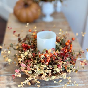 May include: A wooden pedestal with a fall-themed wreath featuring faux leaves and berries. A white pillar candle sits in the center of the wreath.