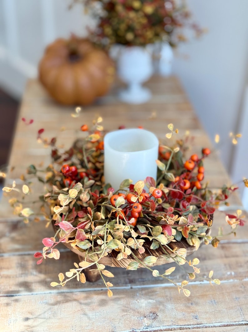 May include: A wooden pedestal with a fall-themed wreath featuring faux leaves and berries. A white pillar candle sits in the center of the wreath.