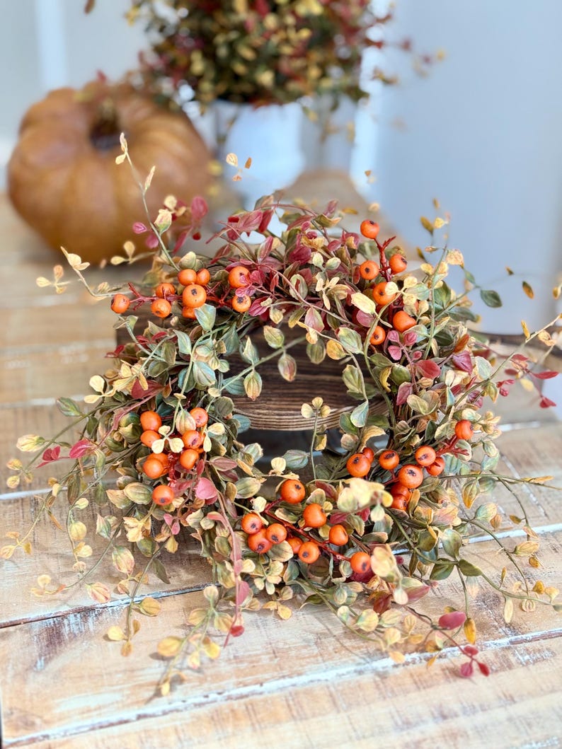May include: A fall-themed wreath with orange berries and green and red leaves. The wreath is made of faux foliage and is perfect for decorating your home for the autumn season.