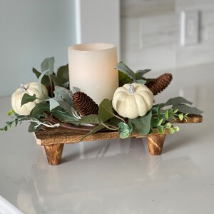 May include: A wooden candle holder with three legs, decorated with faux greenery, white pumpkins, and a pine cone. A white pillar candle sits in the center of the holder.