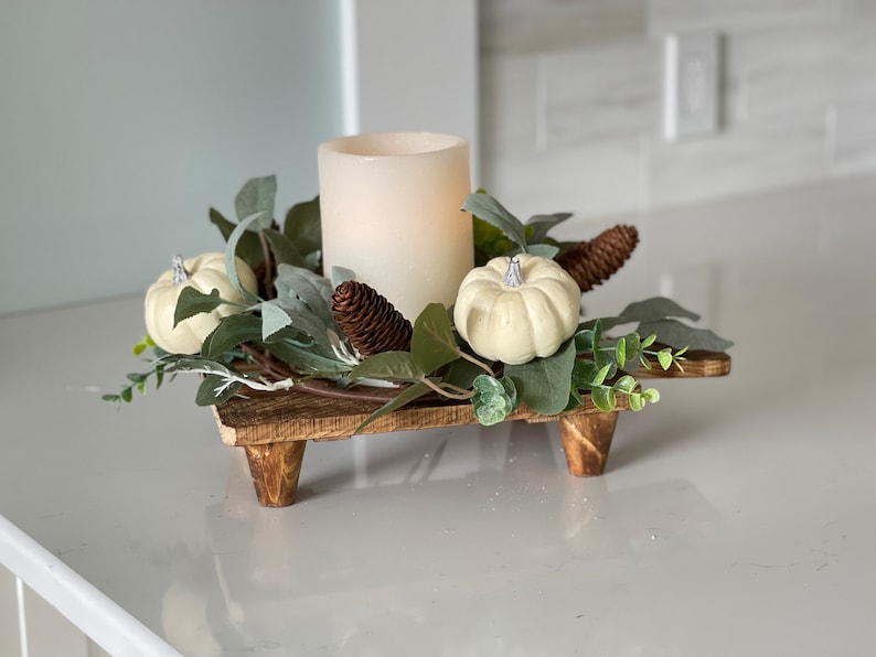 May include: A wooden candle holder with three legs, decorated with faux greenery, white pumpkins, and a pine cone. A white pillar candle sits in the center of the holder.