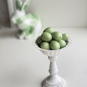 May include: A white pedestal stand with a scalloped edge holds a group of green speckled Easter eggs.