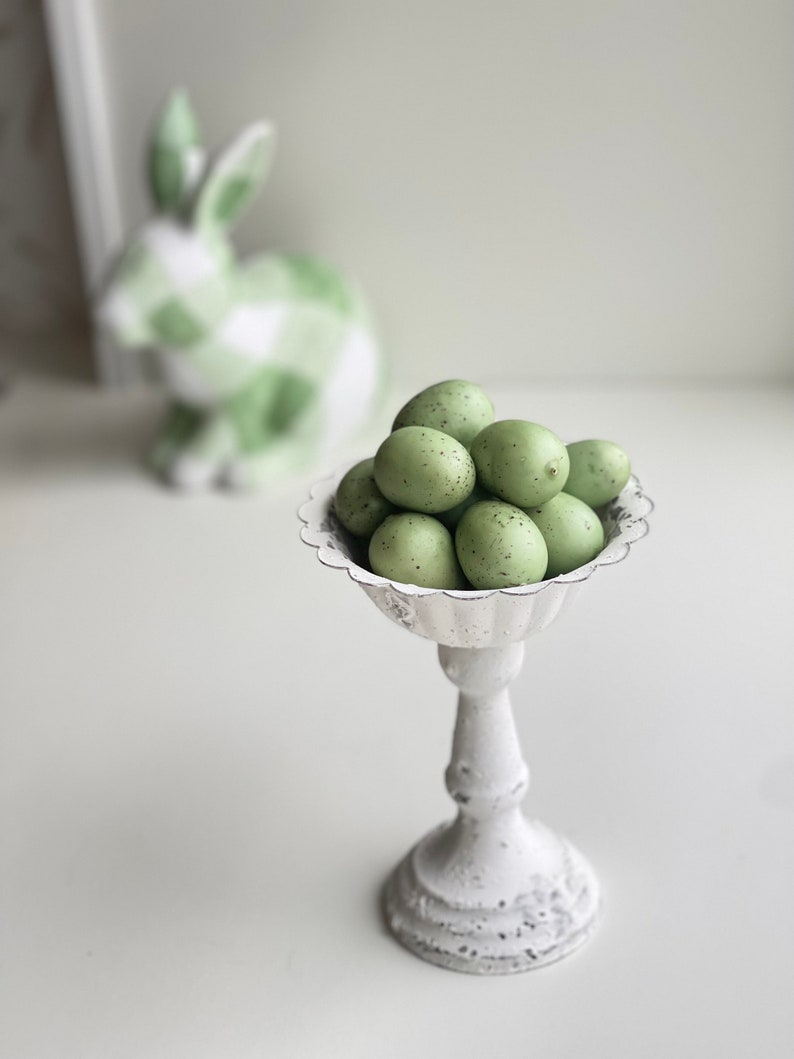 May include: A white pedestal stand with a scalloped edge holds a group of green speckled Easter eggs.
