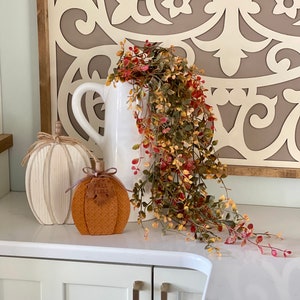 Puede incluir: Una jarra de cerámica blanca con asa está adornada con vegetación en cascada. Una calabaza decorativa con el texto "FALL FEELS" y una calabaza de madera se exhiben sobre una superficie blanca, creando un arreglo temático otoñal.