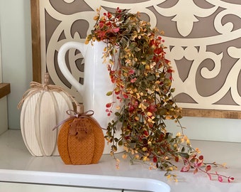 May include: A white ceramic pitcher with a handle is adorned with cascading greenery. A decorative pumpkin with the text "FALL FEELS" and a wooden pumpkin are displayed on a white surface, creating a fall-themed arrangement.