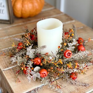 May include: A white pillar candle sits in the center of a fall-themed wreath with red berries, orange and yellow accents, and green foliage. A small framed sign with the words "Hello Fall" is visible.