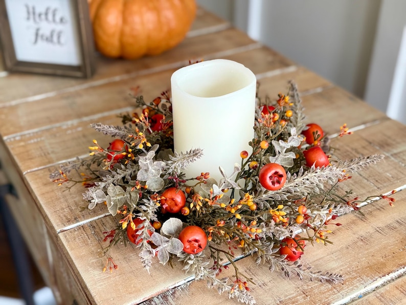 May include: A white pillar candle sits in the center of a fall-themed wreath with red berries, orange and yellow accents, and green foliage. A small framed sign with the words "Hello Fall" is visible.