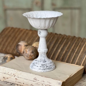 May include: White metal pedestal with a fluted bowl on top. The pedestal is distressed and has a rustic look. It is sitting on an old book with handwritten text.