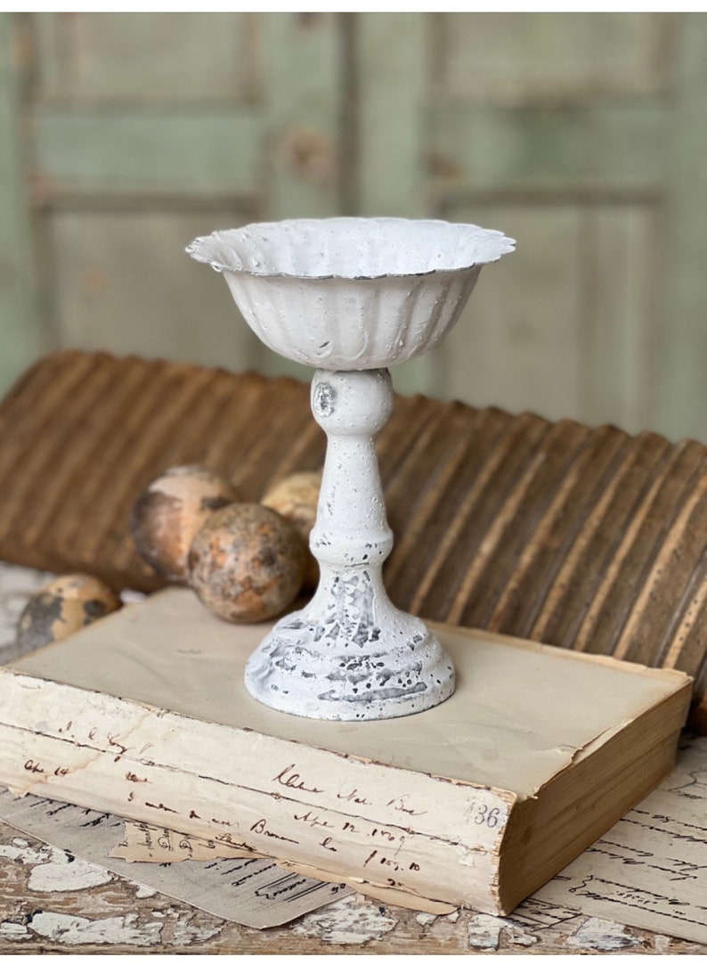 May include: White metal pedestal with a fluted bowl on top. The pedestal is distressed and has a rustic look. It is sitting on an old book with handwritten text.