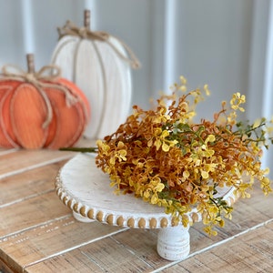 May include: A white wooden pedestal with a beaded edge and three legs. The pedestal is decorated with a bouquet of faux fall foliage in shades of yellow and orange.