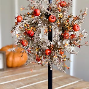 May include: A decorative fall wreath featuring red apple accents, orange berries, and muted green foliage. The wreath is mounted on a black metal stand, creating a seasonal centerpiece. A pumpkin is visible in the background.