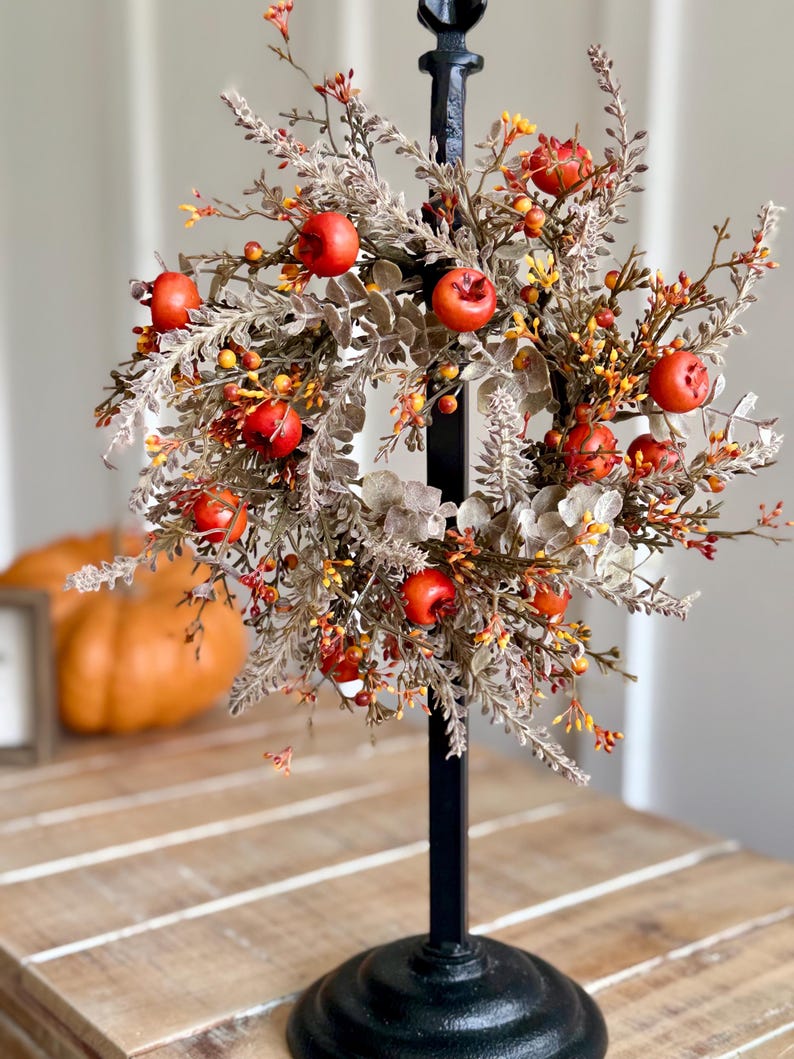 May include: A decorative fall wreath featuring red apple accents, orange berries, and muted green foliage. The wreath is mounted on a black metal stand, creating a seasonal centerpiece. A pumpkin is visible in the background.