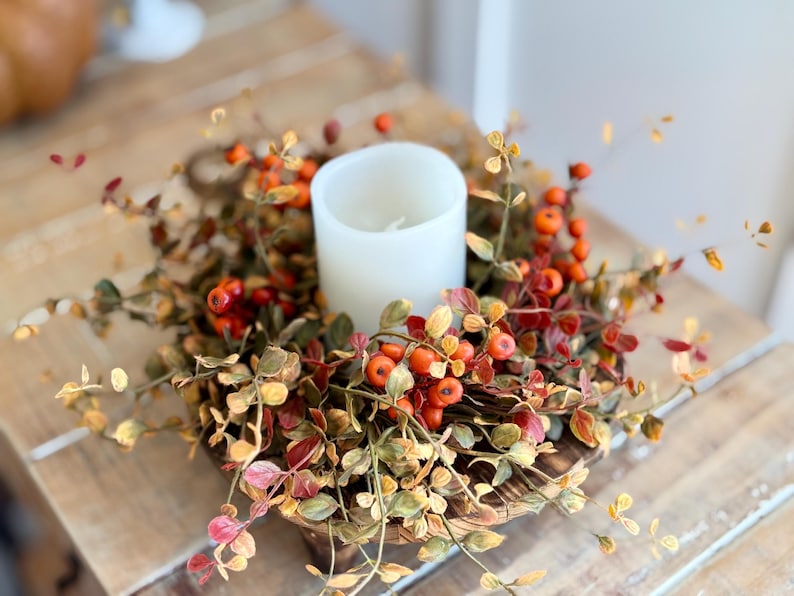 May include: A white pillar candle sits in the center of a fall-themed wreath. The wreath is made of faux greenery with small orange and red berries.