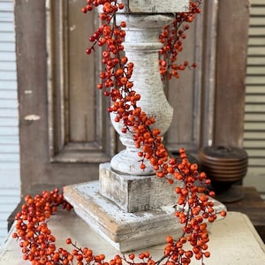 Può includere: Una lunga ghirlanda flessibile caratterizzata da numerose piccole bacche rotonde di colore arancione-rosso brillante attaccate a sottili steli marrone scuro. La ghirlanda è drappeggiata su un piedistallo in legno verniciato di bianco e usurato.