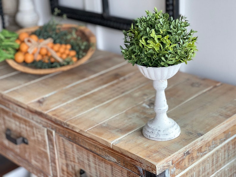 May include: A white pedestal with a green topiary plant on top. The pedestal is on a wooden table with drawers.