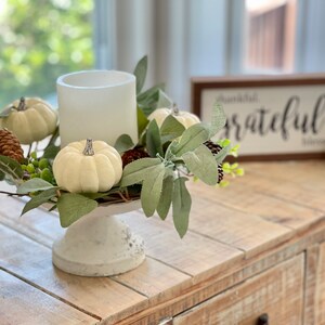 May include: White pumpkins and pine cones arranged in a wreath on a white pedestal with a white candle in the center. The wreath is on a wooden table with a sign that says "thankful" in black lettering.