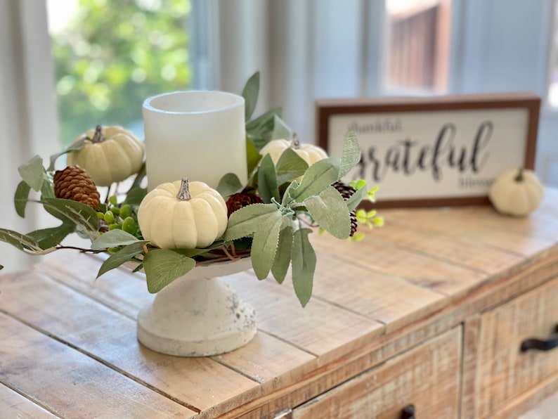 May include: White pumpkins and pine cones arranged in a wreath on a white pedestal with a white candle in the center. The wreath is on a wooden table with a sign that says "thankful" in black lettering.