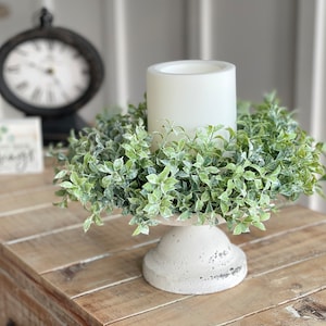 May include: A white pillar candle sits on a concrete pedestal surrounded by a green faux boxwood wreath.