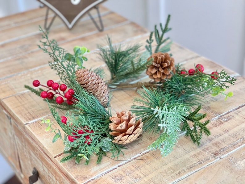 May include: A green pine and cedar Christmas wreath with red berries and brown pine cones. The wreath is on a wooden surface.