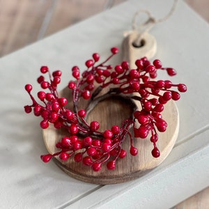 May include: A red berry wreath on a round wooden chopping board. The wreath is made of artificial berries and is ideal for adding a touch of festive cheer to your home.