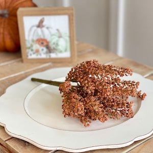 May include: A white charger plate with a scalloped edge, featuring a bouquet of dried brown flowers with a green stem.