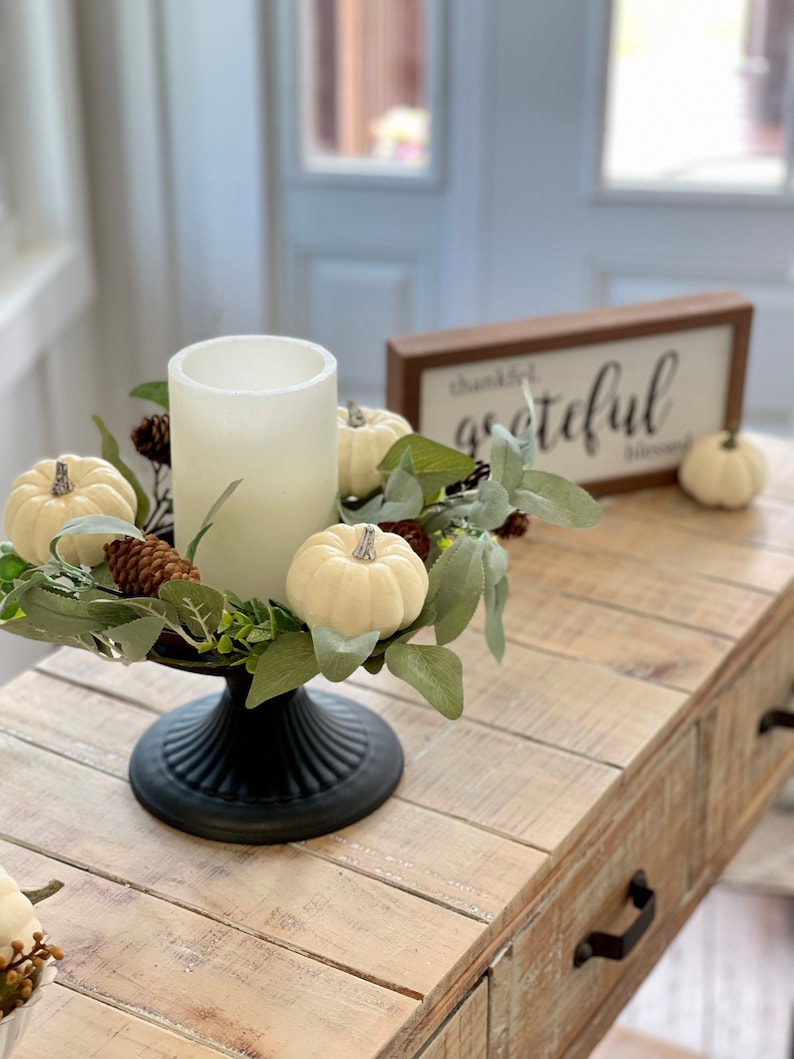 May include: A white pillar candle sits in a black pedestal stand surrounded by a wreath of white pumpkins, pine cones, and eucalyptus leaves. The sign behind the wreath reads "thankful grateful blessed".