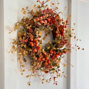 May include: A fall-themed wreath with orange berries and green and yellow leaves. The wreath is made of artificial materials and is perfect for decorating a home for the fall season.