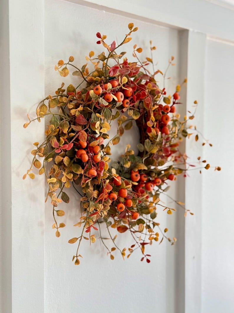 May include: A fall-themed wreath with orange berries and green and yellow leaves. The wreath is made of artificial materials and is perfect for decorating a home for the fall season.