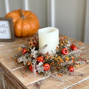 May include: A white candle sits in a decorative fall wreath with red berries and orange accents. A small framed sign with "Hello Fall" is to the left, and a large orange pumpkin is in the background.