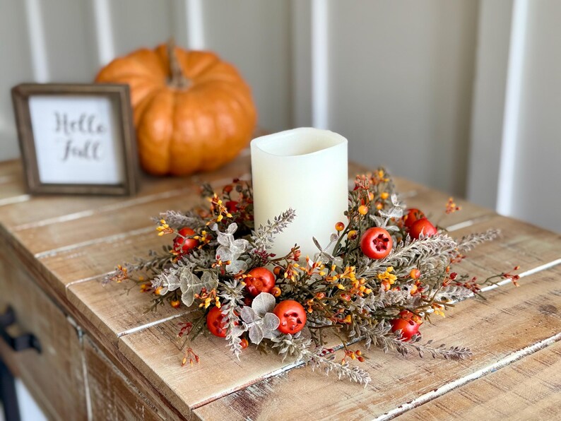 May include: A white candle sits in a decorative fall wreath with red berries and orange accents. A small framed sign with "Hello Fall" is to the left, and a large orange pumpkin is in the background.
