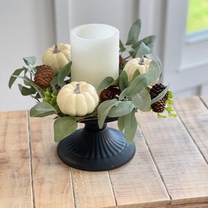 May include: A white pillar candle sits in the center of a black pedestal. The candle is surrounded by a ring of faux greenery, white pumpkins, and pine cones.