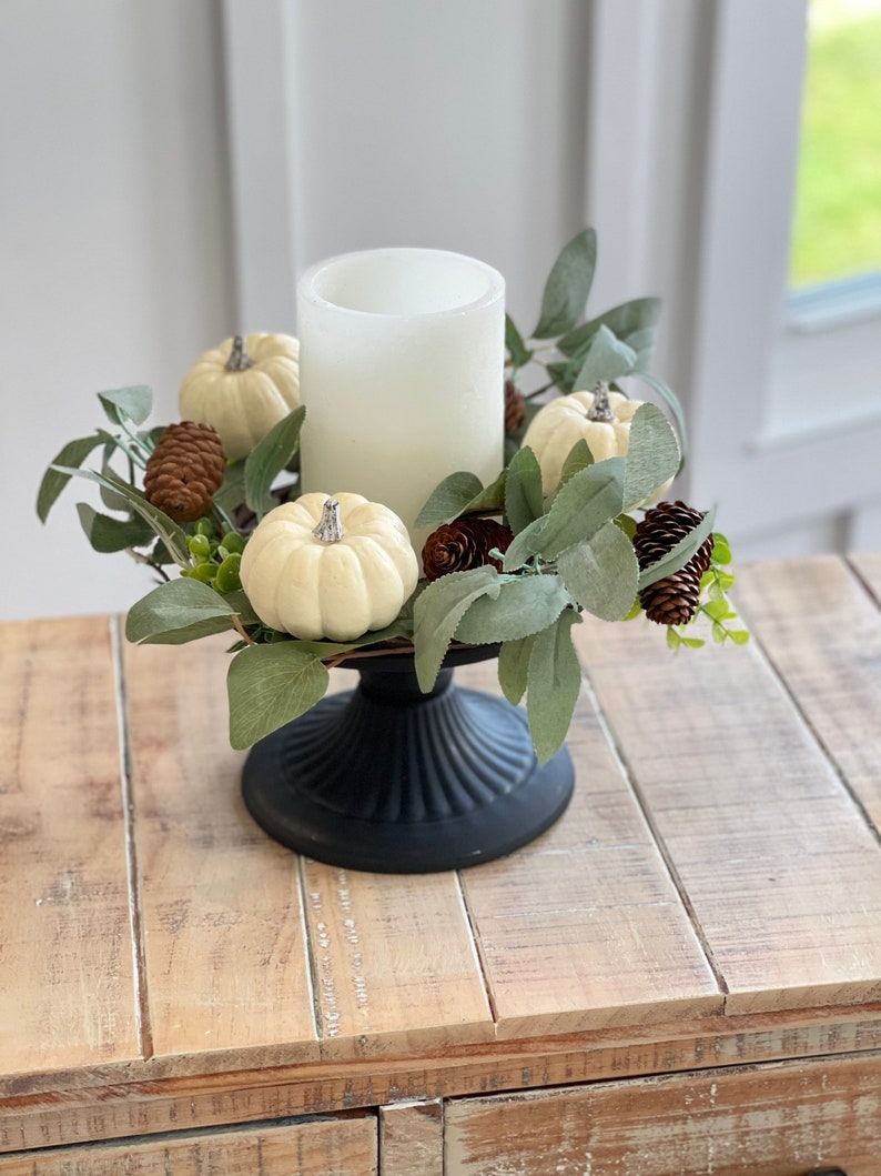 May include: A white pillar candle sits in the center of a black pedestal. The candle is surrounded by a ring of faux greenery, white pumpkins, and pine cones.