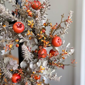 May include: A close-up of a decorative fall arrangement featuring faux red apples, orange berries, and muted green and gray foliage. The arrangement is set against a blurred background, highlighting the autumnal colors and textures.