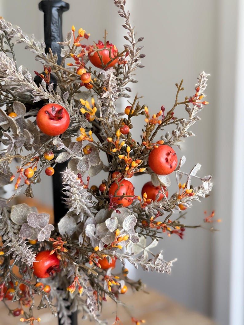 May include: A close-up of a decorative fall arrangement featuring faux red apples, orange berries, and muted green and gray foliage. The arrangement is set against a blurred background, highlighting the autumnal colors and textures.