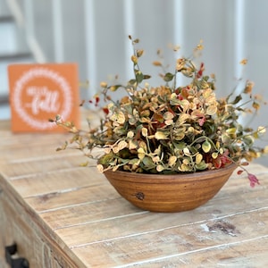 Op de afbeelding: Een houten tafel met een bruine kom gevuld met een bladplant met gele en rode bladeren. Een klein oranje bord met het woord "herfst" in witte letters staat op de tafel.