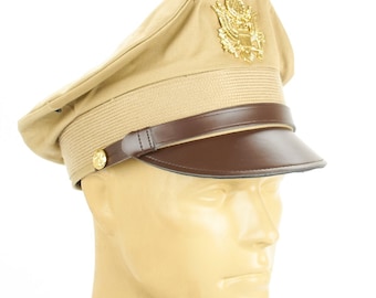 May include: Tan military-style hat with a brown leather visor and band. The hat features a gold emblem and a gold button on the side. The hat is a classic military accessory.