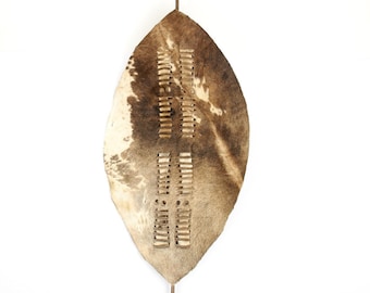 May include: A traditional African shield made of cowhide with a brown and white pattern. The shield is decorated with a series of leather strips and has a metal handle.
