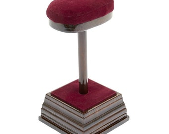 May include: A wooden display stand with a burgundy velvet top and base. The stand has a dark brown, polished finish. The top is oval-shaped, and the base is square with layered edges. The stand is designed to hold a hat or other headwear.