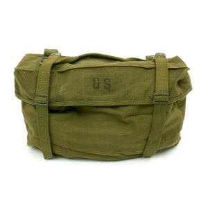May include: Olive green canvas bag with two adjustable straps and a top handle. The bag features the letters "US" printed on the front. The bag is rectangular and appears to be made of durable material, suitable for carrying items. The bag is likely a military-style bag.