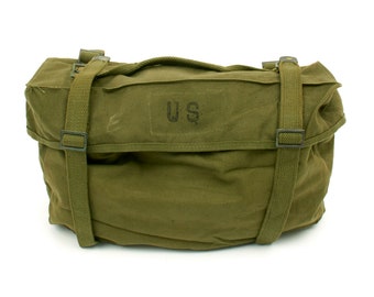 May include: Olive green canvas bag with two adjustable straps and a top handle. The bag features the letters "US" printed on the front. The bag is rectangular and appears to be made of durable material, suitable for carrying items. The bag is likely a military-style bag.