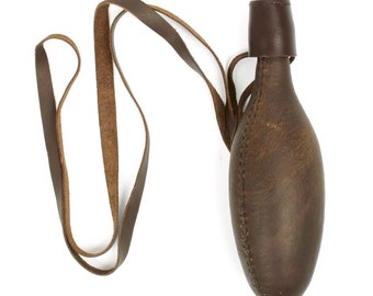 May include: A brown leather flask with a matching leather strap. The flask has a rounded, elongated shape and a small, cylindrical neck. The strap is attached to the neck and extends to form a loop for carrying. The leather appears worn.
